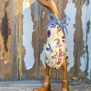 K&ouml;nnte beinhalten: Eine h&ouml;lzerne Entenfigur mit einem floral gemusterten Stoffk&ouml;rper und einem blauen Band um den Hals. Die Ente hat h&ouml;lzerne Beine und Stiefel.