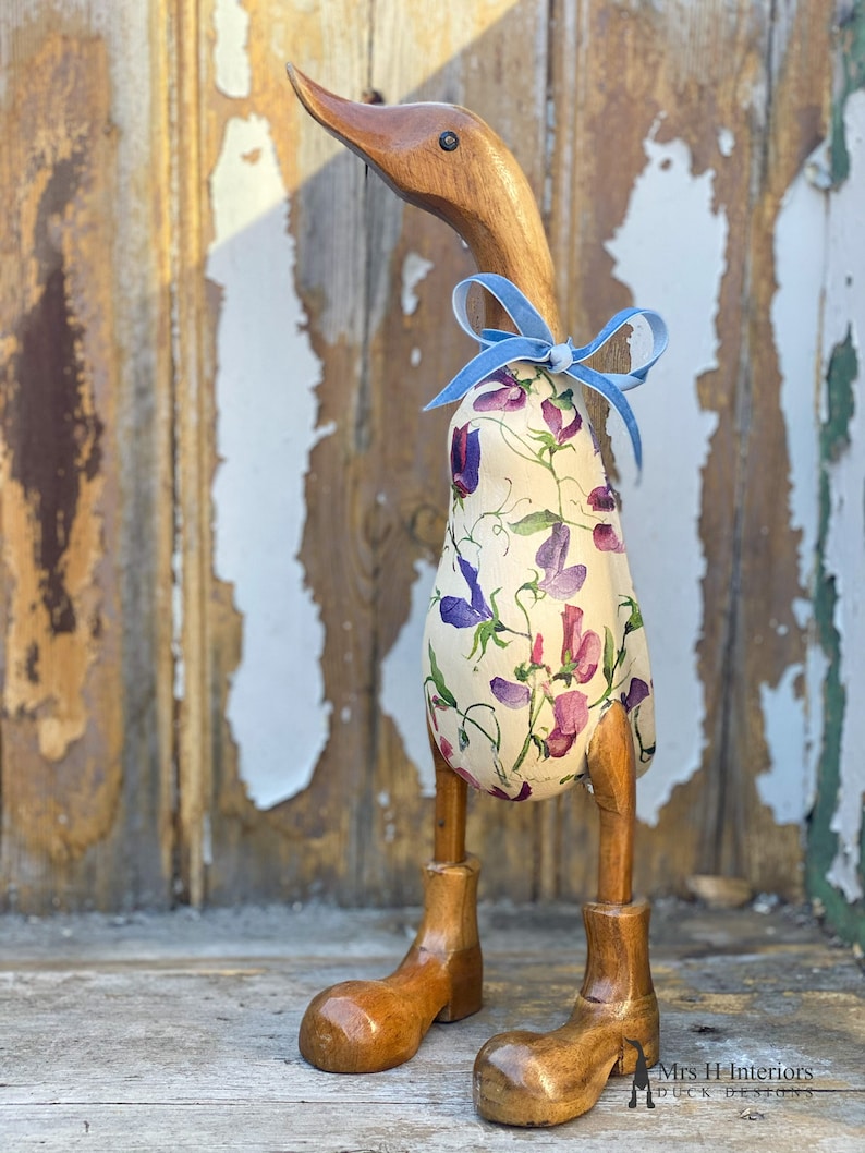K&ouml;nnte beinhalten: Eine h&ouml;lzerne Entenfigur mit einem floral gemusterten Stoffk&ouml;rper und einem blauen Band um den Hals. Die Ente hat h&ouml;lzerne Beine und Stiefel.