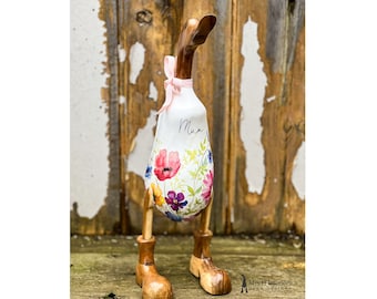 May include: A white wooden duck with floral print and a pink ribbon around its neck. The duck has wooden legs and feet. The duck is standing on a wooden surface. The duck is labeled "Mum".