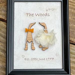 May include: A framed wooden sign with a white background and the text "The Woods" in brown lettering. Two wooden ducks are attached to the sign, one with a yellow bow tie and the other with a white flower. The text "Est. 25th June 1977" is also written on the sign.