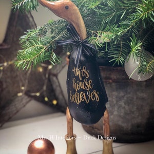 May include: Black wooden duck figurine with gold lettering that reads "this house believes". The duck is wearing a black and white ribbon around its neck and has gold boots.