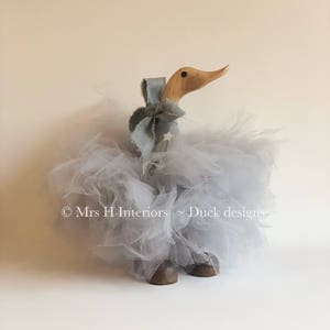 May include: A wooden duck figurine with a gray tulle skirt and a light blue bow. The duck is standing on a white background. © Mrs H Interiors - Duck designs
