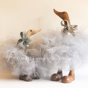 May include: Two wooden duck figurines wearing gray tulle tutus with a light blue bow on each. The larger duck has a gold ribbon around its neck. The text "Mrs H Interiors - Duck designs" is visible in the bottom center of the image.