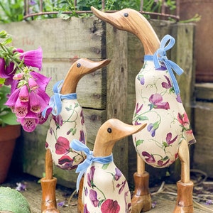 K&ouml;nnte beinhalten: Drei h&ouml;lzerne Enten, die mit einem Blumenmuster aus rosa und violetten Blumen verziert sind. Die Enten tragen blaue Samtband-Schleifen um den Hals. Die Enten stehen auf einer Holzoberfl&auml;che. Der Text "Mrs H Interiors Duck Designs" ist in der rechten unteren Ecke des Bildes sichtbar.