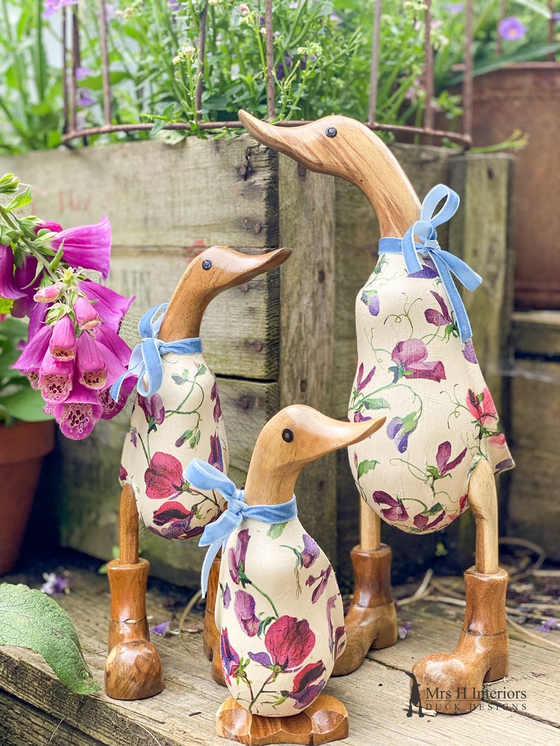 K&ouml;nnte beinhalten: Drei h&ouml;lzerne Enten, die mit einem Blumenmuster aus rosa und violetten Blumen verziert sind. Die Enten tragen blaue Samtband-Schleifen um den Hals. Die Enten stehen auf einer Holzoberfl&auml;che. Der Text "Mrs H Interiors Duck Designs" ist in der rechten unteren Ecke des Bildes sichtbar.