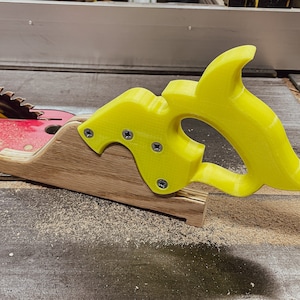 Könnte beinhalten: Ein gelber Kunststoff-Jig mit Griff, der an einer Holzbasis befestigt ist, zur Verwendung mit einer Tischkreissäge.