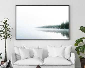 May include: A white sofa with two pillows sits in a living room with a black and white rug. A coffee table with a copper top sits in front of the sofa. A framed photograph of a foggy lake with trees in the background hangs on the wall above the sofa.
