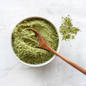 May include: A white bowl filled with green matcha powder and a wooden spoon. Some matcha powder is scattered on the white surface.