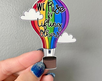 May include: A rainbow hot air balloon with the text "We Rise By Lifting Others" in black lettering. The balloon has white clouds on either side.
