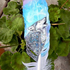 Puede incluir: Una pluma blanca con un dise&ntilde;o pintado de un lobo aullando a la luna. La pluma est&aacute; pintada en tonos de azul, morado y blanco. El lobo est&aacute; pintado en tonos de gris y blanco.