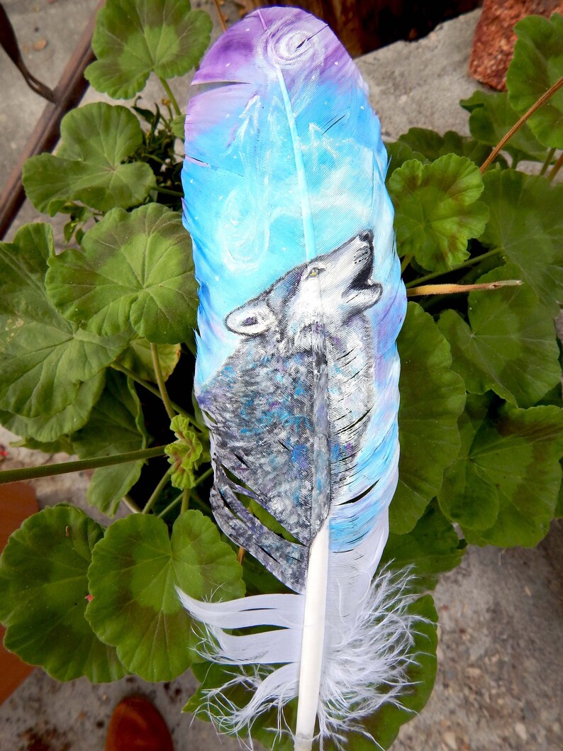 Puede incluir: Una pluma blanca con un dise&ntilde;o pintado de un lobo aullando a la luna. La pluma est&aacute; pintada en tonos de azul, morado y blanco. El lobo est&aacute; pintado en tonos de gris y blanco.
