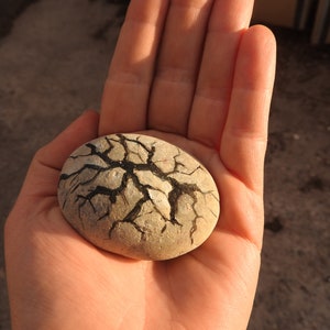 May include: A smooth, round, light grey stone with black painted cracks. The stone is being held in a person's hand.