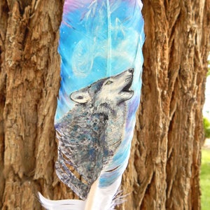 Puede incluir: Una pluma pintada a mano con un lobo aullando a la luna. La pluma est&aacute; pintada en tonos de azul, morado y blanco, con un dise&ntilde;o en espiral. El lobo est&aacute; pintado en tonos de gris y blanco.