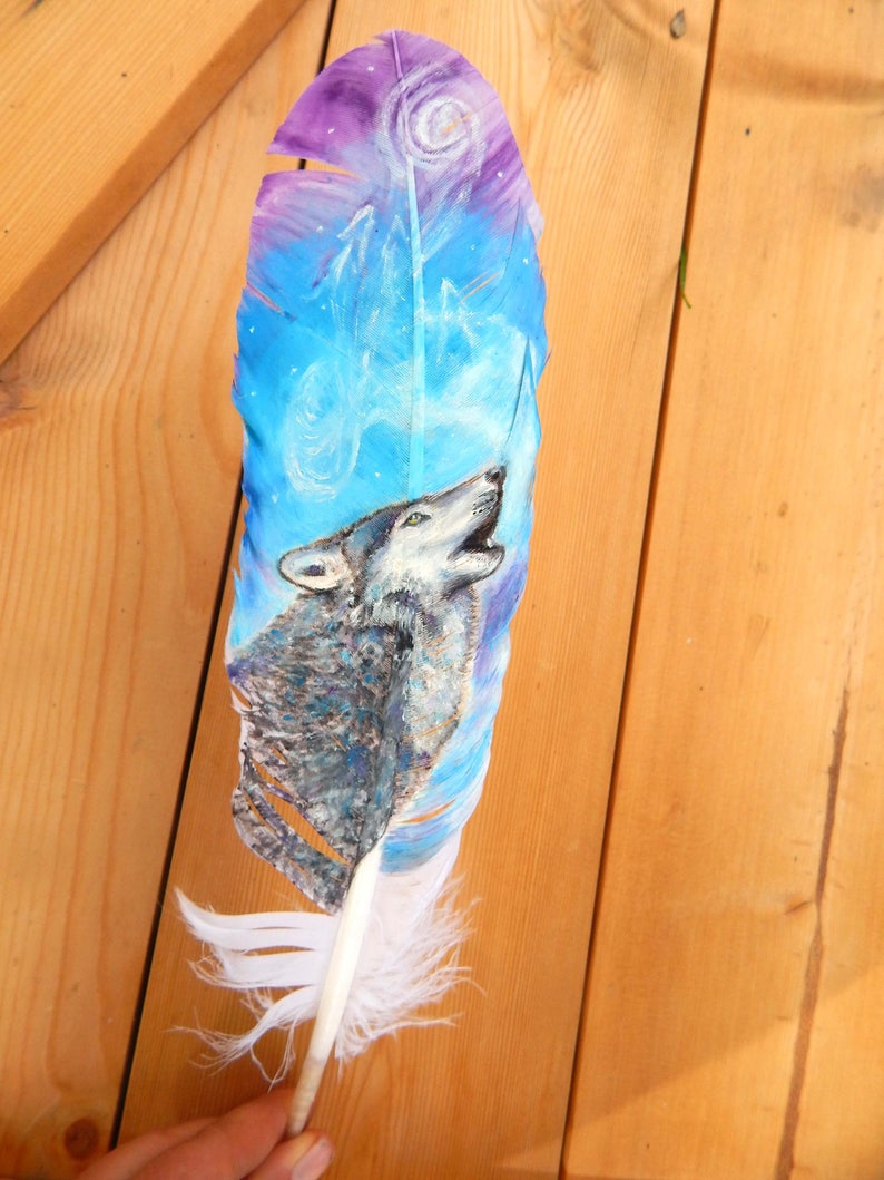 Puede incluir: Una pluma pintada a mano con un lobo aullando a la luna. La pluma est&aacute; pintada en tonos de azul, morado y blanco, con un dise&ntilde;o en espiral en el fondo. El lobo est&aacute; pintado en tonos de gris y blanco.