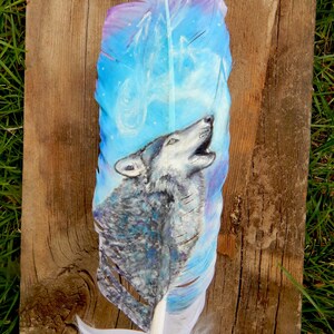 Puede incluir: Una pluma pintada con un lobo aullando a la luna. La pluma est&aacute; pintada en tonos de azul, morado y blanco, con una luna blanca y estrellas. El lobo est&aacute; pintado en tonos de gris y blanco.