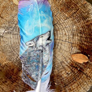 Puede incluir: Una pluma blanca con un dise&ntilde;o pintado de un lobo aullando a la luna. El lobo est&aacute; pintado en tonos grises y azules, y la luna est&aacute; pintada en tonos morados y azules. La pluma est&aacute; sobre un fondo de madera.