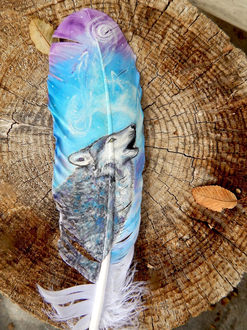 Puede incluir: Una pluma blanca con un dise&ntilde;o pintado de un lobo aullando a la luna. El lobo est&aacute; pintado en tonos grises y azules, y la luna est&aacute; pintada en tonos morados y azules. La pluma est&aacute; sobre un fondo de madera.