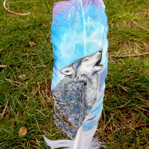 Puede incluir: Una pluma blanca con un dise&ntilde;o pintado de un lobo aullando a la luna. El lobo es gris y blanco, y la luna est&aacute; rodeada de un cielo azul y morado. La pluma es una hermosa obra de arte.