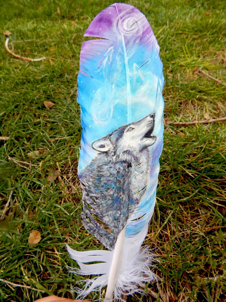 Puede incluir: Una pluma blanca con un dise&ntilde;o pintado de un lobo aullando a la luna. El lobo es gris y blanco, y la luna est&aacute; rodeada de un cielo azul y morado. La pluma es una hermosa obra de arte.