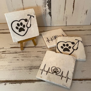 May include: Set of four white ceramic coasters with black paw print designs. One coaster features a paw print inside a heart with a stethoscope. Two coasters feature a paw print inside a heart with a heartbeat line. One coaster is on a small wooden easel.