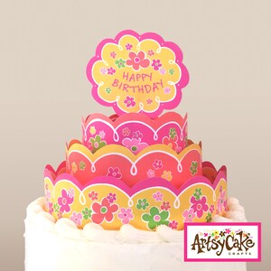 Może przedstawiać: Trzypiętrowy tort z kwiatowym wzorem. Ciasto jest ozdobione różowymi, żółtymi i zielonymi wzorami kwiatowymi. Na wierzchu ciasta znajduje się topper "Happy Birthday". Ciasto ma białą podstawę z lukru.