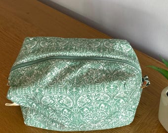 May include: A rectangular cosmetic bag with a green and white floral pattern and a green zip. The bag is on a wooden surface, next to a small potted plant with green leaves.