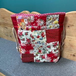 Puede incluir: Una bolsa con cordón hecha de un mosaico de varias telas con patrones rojos, blancos y florales. El borde superior está acabado con un ribete de cuadros vichy rojo y blanco, y la bolsa incluye cordones gruesos de cuerda trenzada color crema.