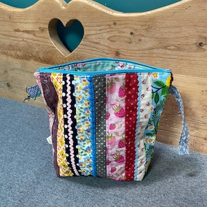 May include: A quilted fabric pouch featuring vertical strips of various colorful patterns, including strawberries, floral prints, and polka dots. The bag has a bright blue zipper closure at the top and two side attachment loops for a strap.