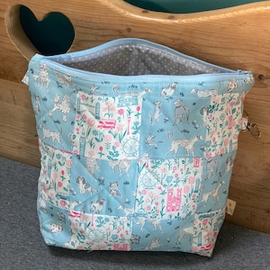 May include: A light blue quilted tote bag with a zip closure. The bag displays a repeating pattern of dogs, flowers, and architectural elements in white, pink, and teal. The interior lining is light grey with white polka dots.