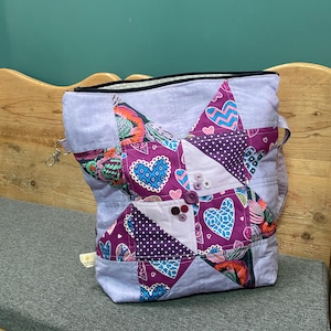 Può includere: Una borsa tote fatta a mano con un design patchwork a forma di stella. La borsa presenta uno sfondo azzurro con punte di stella viola decorate con motivi a cuore e pois. Ha una cerniera nera e una piccola tasca con bottoni.