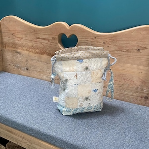 Peut inclure: Un sac à cordon patchwork aux tons bleu clair et crème. Le sac présente divers motifs de tissu, notamment des imprimés floraux et journaux. Le sac est posé sur un coussin bleu.