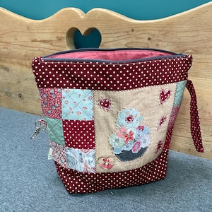 May include: A handmade patchwork zippered pouch with a floral design. The pouch features a burgundy polka dot fabric, a beige centre panel with a floral applique, and various patterned fabric squares. A small key ring is attached.