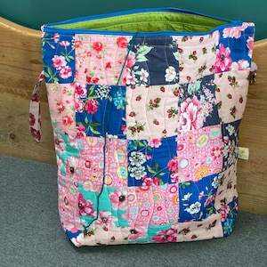 May include: A quilted tote bag with a patchwork design. The bag features squares of pink, blue, and navy fabric, each adorned with floral patterns. The interior is lined with green fabric, and the bag has a blue zipper and handles.