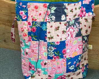 May include: A patchwork tote bag with a floral design. The bag is made from squares of pink, blue, and navy fabric, each with a different floral pattern. The interior is lined with green fabric, and it has a blue zip and handles.