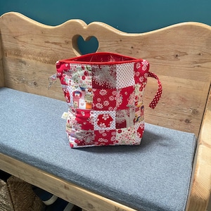 May include: A patchwork fabric tote bag with a red interior and handles. The bag features a variety of red and white floral and patterned squares. The bag is sitting on a gray cushion.