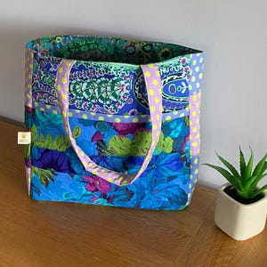 May include: A patterned tote bag featuring a vibrant mix of blue, purple, and green floral designs. The handles are light purple with yellow polka dots. The bag's interior is green with a floral pattern. It sits on a wooden surface.