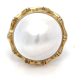 May include: A gold-toned ring with a large, round, white pearl center. The ring's band has a textured, bamboo-like design. The pearl is smooth and lustrous, set against the warm gold.