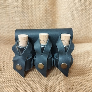 Może przedstawiać: Trzy małe szklane butelki z korkowymi korkami są trzymane w czarnym skórzanym uchwycie. Uchwyt ma trzy pętle do zabezpieczenia butelek i metalowy guzik na każdej pętli.