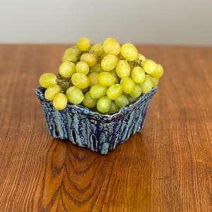 May include: A ceramic berry basket filled with green grapes. The basket is a dark blue with a textured, rustic design. The grapes are a pale green color and are clustered together.