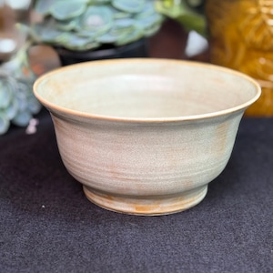 May include: A beige ceramic bowl with a wide, shallow design. The bowl has a slightly flared rim and sits on a small, rounded base. The exterior has a textured appearance with subtle colour variations, suggesting a handmade quality. The bowl is suitable for serving or display.