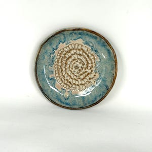 May include: A handmade ceramic plate with a spiral design. The plate features a light blue rim with a brown edge, surrounding a textured, off-white spiral pattern. The center has a light pink hue. This decorative plate is perfect for serving appetizers or as a unique home decor item.