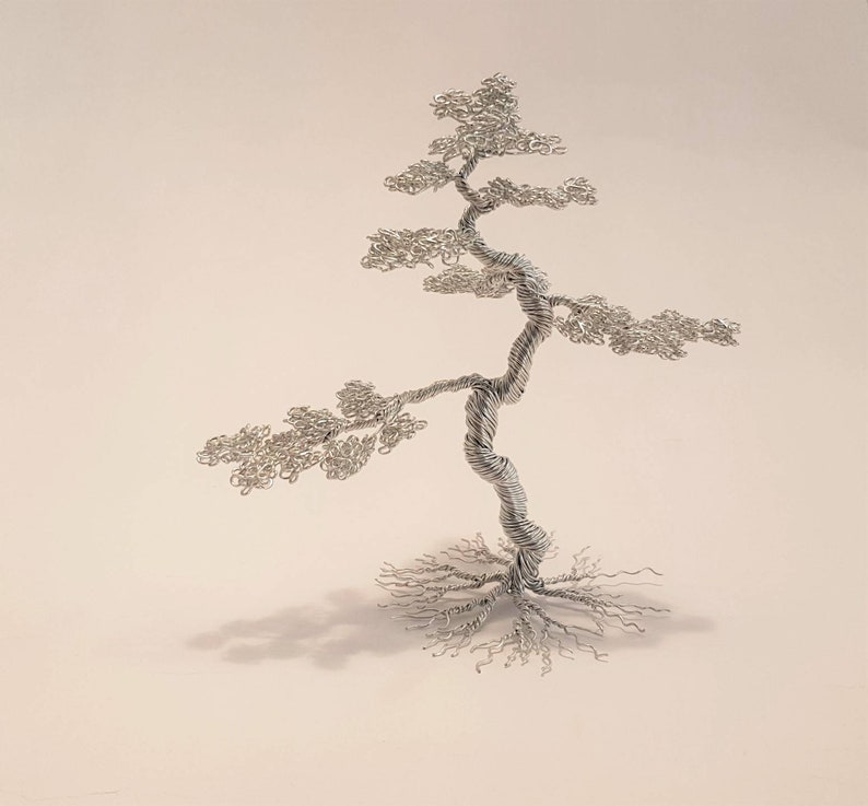 May include: A handcrafted silver wire sculpture of a bonsai tree. The tree features a twisted trunk, delicate branches, and a detailed root system. The sculpture is set against a neutral background, highlighting the intricate details of the wire art.