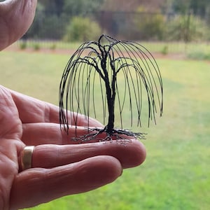 May include: A small, black wire sculpture of a weeping willow tree. The tree is held in a person's hand, with the branches cascading down.
