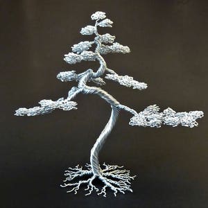 May include: A silver-colored wire sculpture of a bonsai tree against a dark background. The tree features a twisted trunk, detailed branches, and a root system. The leaves are textured, and the overall design is intricate.