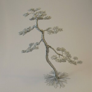 May include: A silver wire sculpture of a bonsai tree in the slanting style, approximately 7 inches tall. The tree has a twisted trunk and delicate, leafy branches. The base of the tree is a network of wire roots. Text on the image reads: Slanting Style - 18 cm. tall.