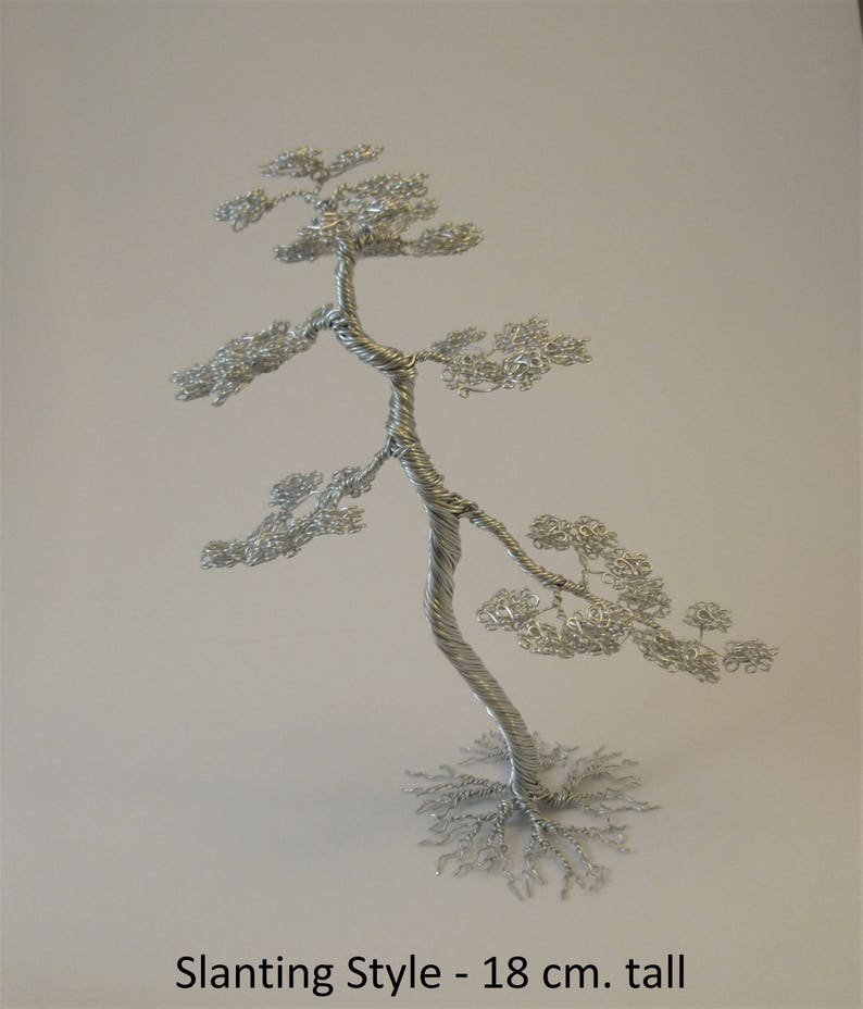 May include: A silver wire sculpture of a bonsai tree in the slanting style, approximately 7 inches tall. The tree has a twisted trunk and delicate, leafy branches. The base of the tree is a network of wire roots. Text on the image reads: Slanting Style - 18 cm. tall.
