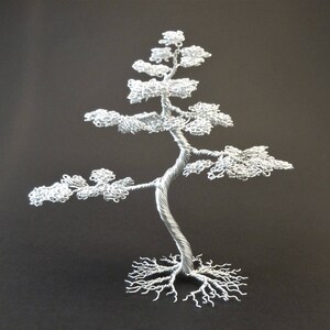 May include: A silver wire sculpture of a bonsai tree. The tree has a twisted trunk, detailed branches, and a full crown of leaves. The roots are also intricately crafted. The sculpture is set against a dark background.