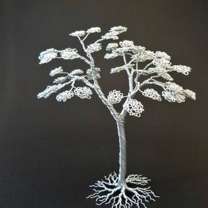 May include: A handcrafted silver wire tree sculpture against a black background. The tree features a detailed trunk, branches, and delicate, textured leaves. The roots are also intricately designed. This unique piece of art is a decorative item.