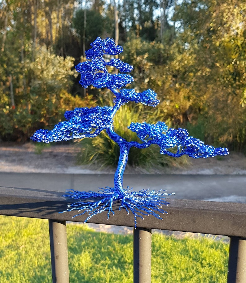 May include: A handcrafted, blue wire bonsai tree sculpture. The tree features a detailed trunk, branches, and foliage, with exposed roots. The sculpture is made of blue wire and sits on a black surface, with a natural background.
