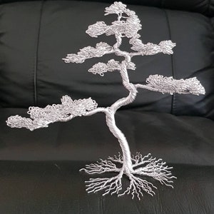 May include: A silver wire sculpture of a bonsai tree. The tree has a twisted trunk, branches with textured foliage, and exposed roots. The sculpture is displayed on a black surface.